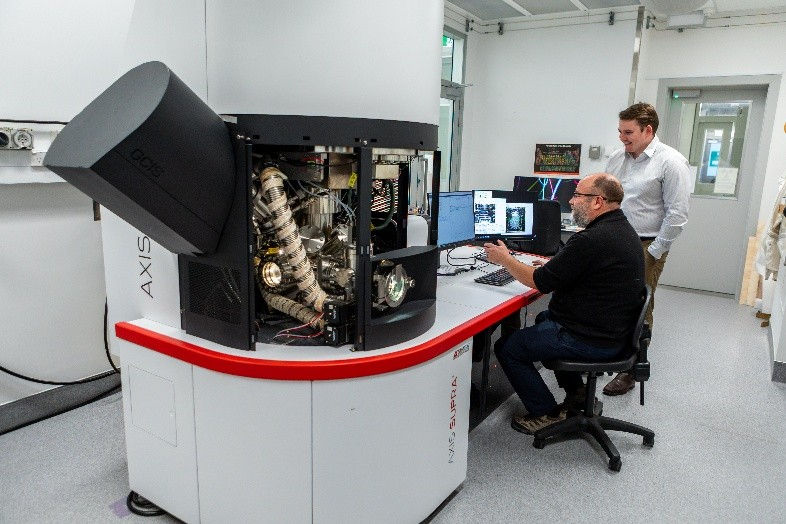 A man sitting at a computer looking at scanned imagery, beside a large x-ray photoelectron spectroscopy machine. Another man peers over his shoulder at the computer screen.